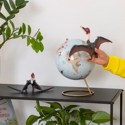 Figuras de dinossauro voador castanho e rosa e globo terrestre azul claro com suporte dourado numa mesa preta com plantas ao fundo