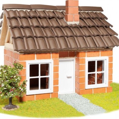 Casa miniatura com paredes de tijolo vermelho, telhado castanho, janelas e porta branca, chaminé, árvore e relva.