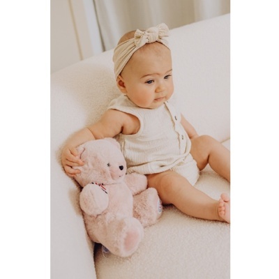 Bebé com roupa branca de malha e faixa de cabeça, sentado num sofá branco com um urso de peluche rosa