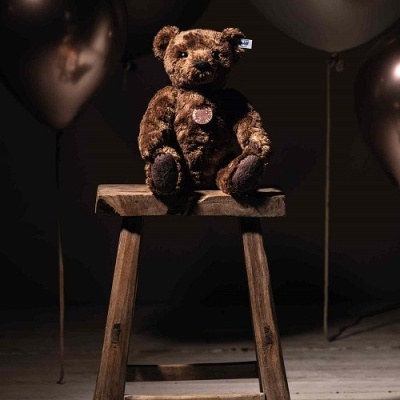 Urso de peluche castanho num banco de madeira com balões pretos ao fundo