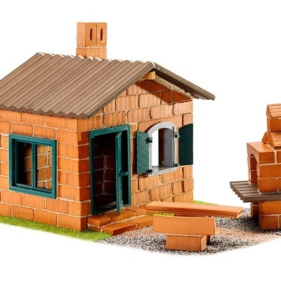 Miniatura de casa em tijolos com porta e janela verdes, telhado cinzento e lareira exterior com mesa e bancos.