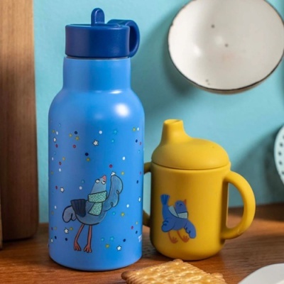 Garrafa azul e copo amarelo com desenhos de pássaros, tampas e pegas, sobre superfície de madeira com bolachas