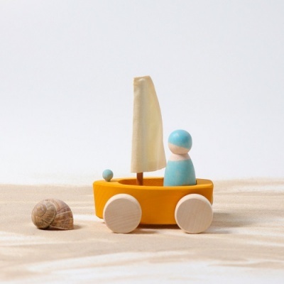 Barco de madeira amarelo com vela creme e figura azul sobre rodas ao lado de concha