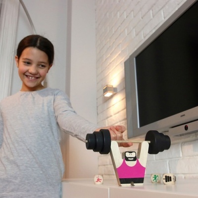 Brinquedo de halterofilista em madeira com menina sorridente ao lado, parede branca de tijolos e televisão