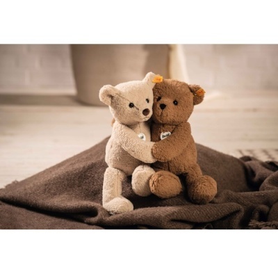 Dois ursos de peluche abraçados sobre cobertor castanho em chão de madeira clara