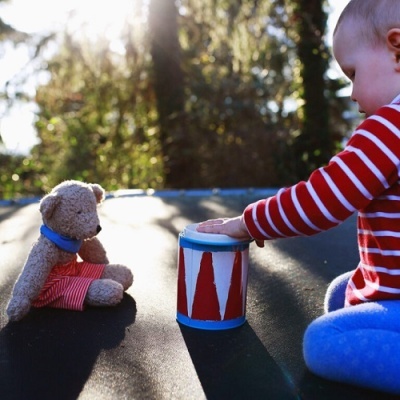 Criança com roupa riscada a tocar num tambor com urso de peluche sentado