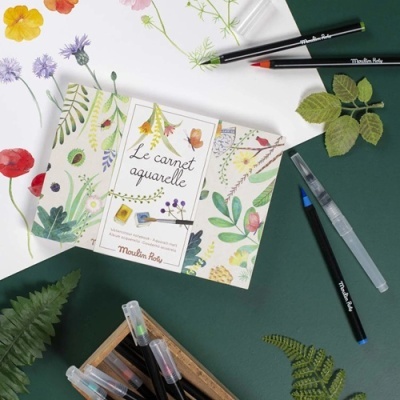 Caderno de aguarela com capa floral da Moulin Roty e pinceis de aguarela sobre fundo verde.