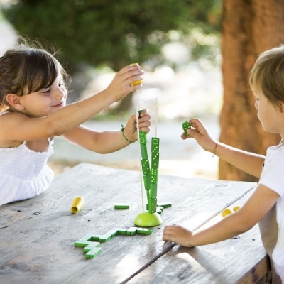 Duas crianças brincando com jogo de empilhar peças verdes numa mesa de madeira ao ar livre.