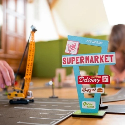 Sinal de supermercado em madeira colorida com textos variados sobre superfície de jogo e modelo de guindaste amarelo ao fundo.