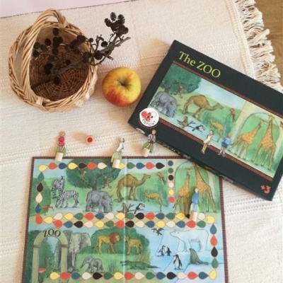Jogo de tabuleiro The ZOO com peças e animais, cesta de vime e maçã numa superfície têxtil clara