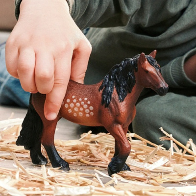 Brinquedo de cavalo castanho com crina preta, manchas brancas, mão de criança a segurar.
