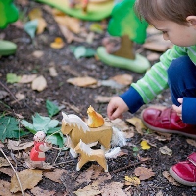 Criança a brincar com brinquedos de madeira de animais e figura humana no chão com folhas