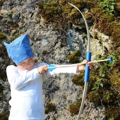 Criança com túnica branca e chapéu azul segurando arco com flecha de ventosa em ambiente rochoso