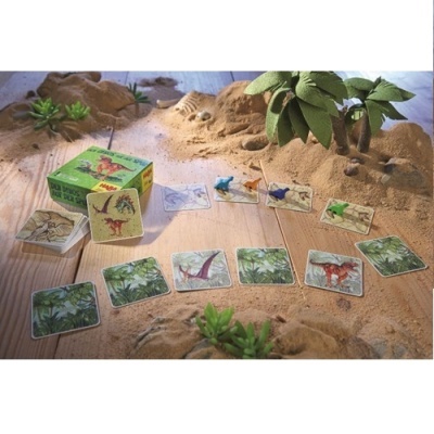 Cartas de jogo com dinossauros coloridos, caixa verde e miniaturas sobre mesa com areia e plantas artificiais