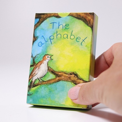 Caixa ilustrada com um pássaro e o texto The alphabet, segurada por uma mão