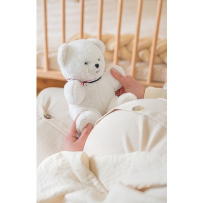 Ursinho de pelúcia branco segurado por pessoa vestida de branco junto a berço de madeira