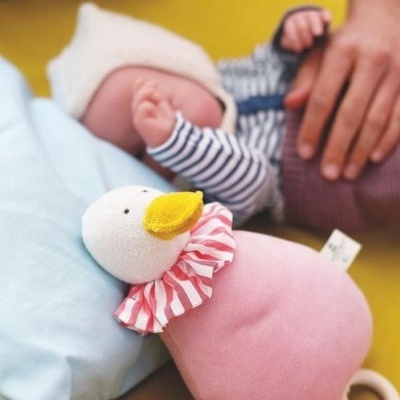 Brinquedo de pato de tecido rosa com bico amarelo e babado às riscas, com bebé e almofada azul no fundo amarelo