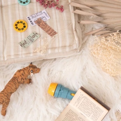 Manta bege com franjas, View-Masters coloridos, figura de tigre, lanterna azul e livro aberto sobre tapete branco felpudo