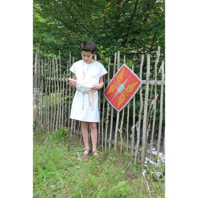 Menino com túnica branca e sandálias lê livro junto a cerca rústica com bandeira vermelha e amarelo