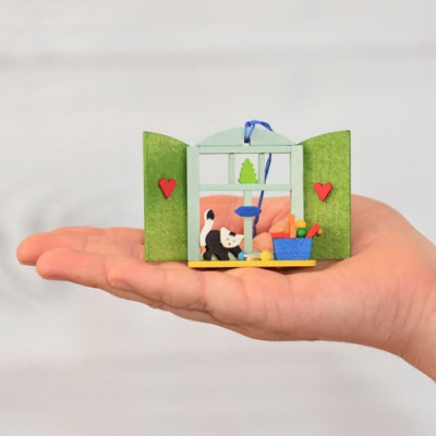 Miniatura de janela verde com gato, árvore e cesta azul sendo segurada numa mão