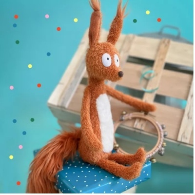 Boneco de pelúcia castanho com barriga branca sentado num presente azul, fundo azul claro com confetis coloridos