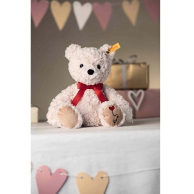 Urso de peluche branco com laço vermelho sentado sobre mesa decorada com bandeirolas e presentes