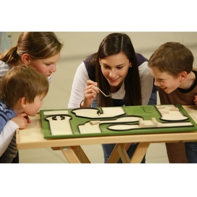 Tabuleiro de madeira com peças recortadas em mesa e quatro pessoas observando.