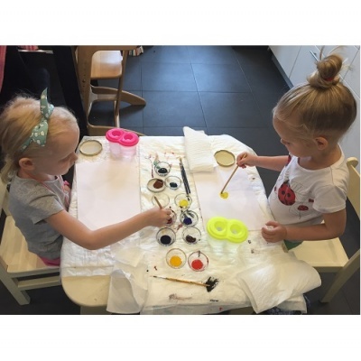 Duas meninas pintam com pincéis e tintas coloridas numa mesa branca