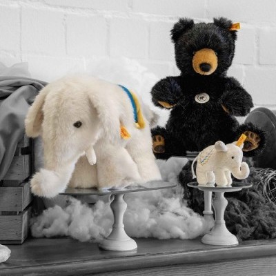 Três peluches: elefante branco, urso preto, elefante branco pequeno, sobre suportes brancos em ambiente com parede branca