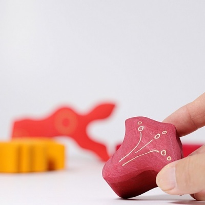 Peça decorativa vermelha de madeira com padrão branco, segurada por mão, com outras peças coloridas desfocadas ao fundo