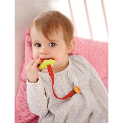 Bebé com vestido cinza claro a morder brinquedo mordedor verde com fita vermelha e clip amarelo numa almofada rosa.