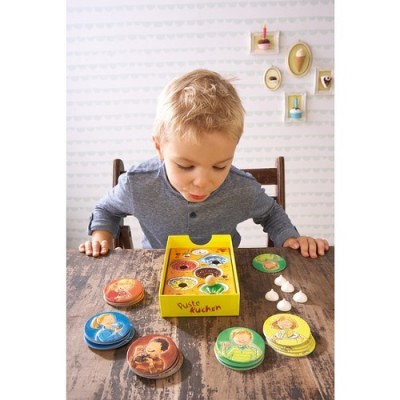 Criança com jogo de tabuleiro com caixa amarela 'Puste Kuchen' sobre mesa de madeira