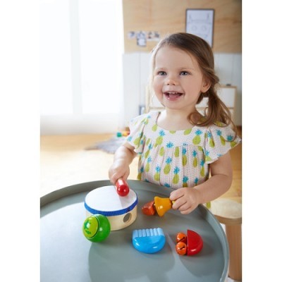 Criança a brincar com tambor de madeira e acessórios coloridos numa mesa cinzenta