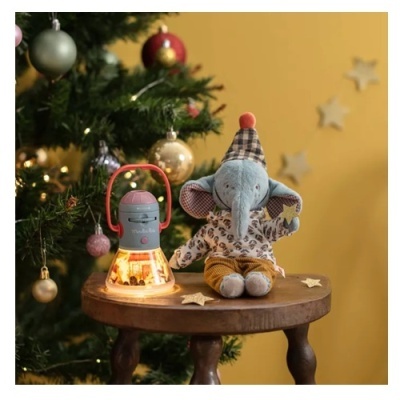 Peluches de elefante azul com chapéu e roupa, lanterna iluminada, mesa de madeira, árvore de Natal decorada, fundo amarelo com estrelas