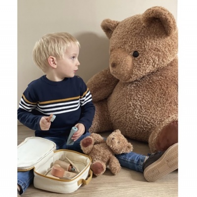 Criança loira sentada junto a urso de peluche grande e brinquedos sobre chão de madeira