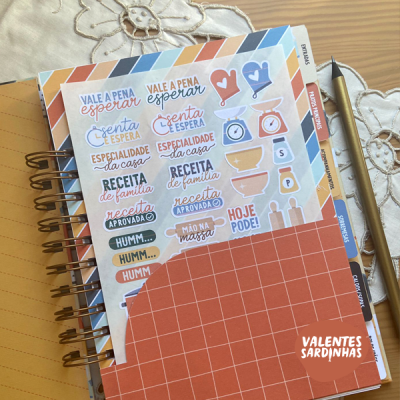 Caderno com capas quadriculadas laranja e branca com texto e etiquetas autocolantes coloridas relacionadas com receitas, numa mesa de madeira com caneta prateada