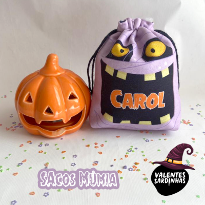 Saco de pano roxo com rosto e nome CAROL ao lado de abóbora laranja decorativa em fundo branco com confetes