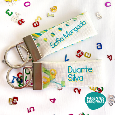 Dois porta-chaves de tecido branco personalizados com nomes, com fecho metálico, em fundo branco com letras e números coloridos