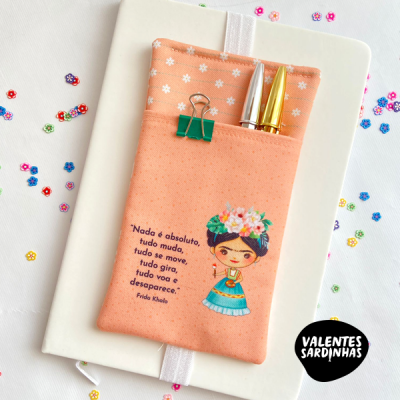 Capa de tecido cor de pêssego com image e texto, clipe verde e canetas douradas sobre caderno branco com lantejoulas coloridas