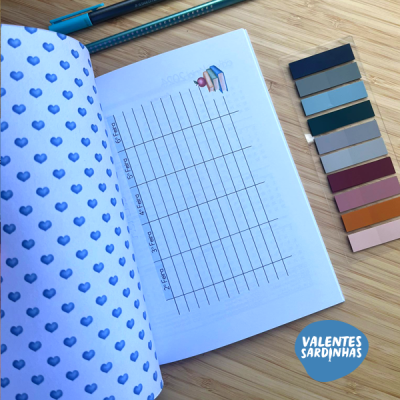 Caderno aberto com padrão de corações azuis, tabela para preenchimento, fitas coloridas e canetas azuis sobre fundo de madeira clara.