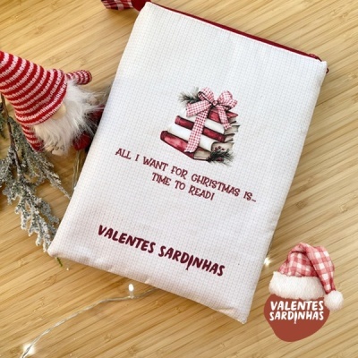 Bolsa branca com fecho vermelho, livros ilustrados com laço e texto natalício, sobre superfície de madeira com decoração de Natal