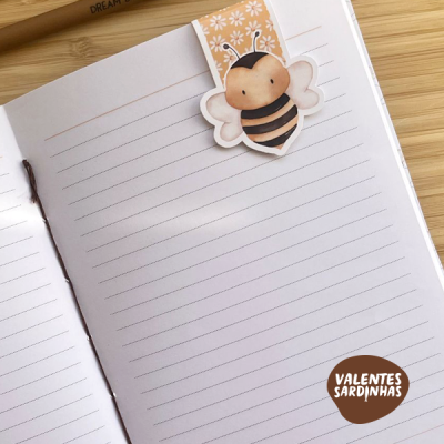 Caderno aberto com fita marcadora de abelha num fundo de madeira clara