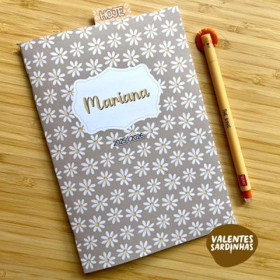 Agenda cinzenta com flores brancas, nome Mariana e ano 2025/2026, com caneta castanha ao lado