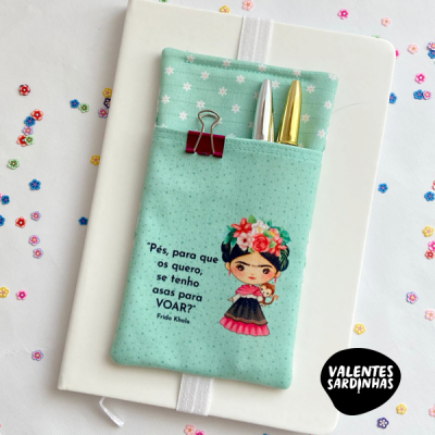 Estojo tecido azul claro com ilustração e texto de Frida Kahlo, canetas e clipe sobre caderno branco