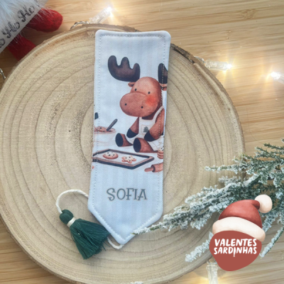 Marcador de livro tecido branco com desenho de alce e nome SOFIA, com franjas verde escuro, sobre base de madeira circular com decoração de Natal