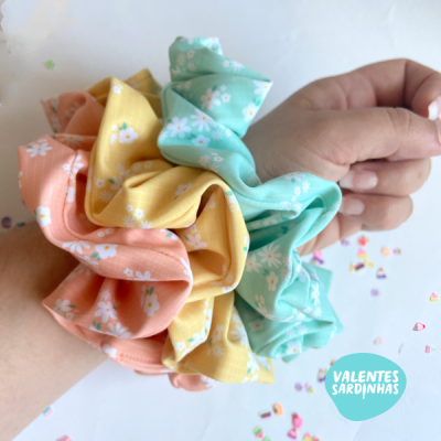 Três scrunchies florais coloridos em dois pulsos com fundo branco e confetes
