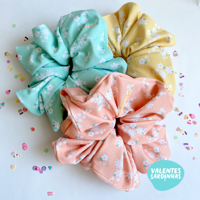 Três scrunchies florais coloridos em verde, amarelo e salmão sobre fundo branco com confetes e logotipo