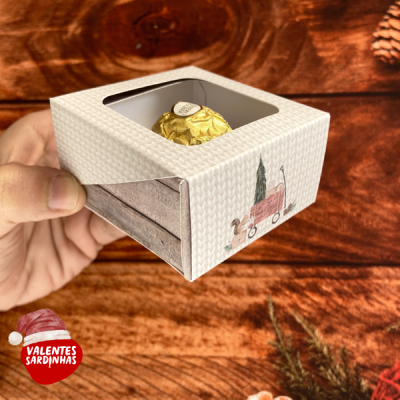 Caixa branca texturizada com bombom dourado vista por janela transparente, decorada com desenho de carrinho vermelho e árvore.