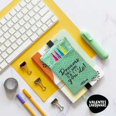 Estojo azul-menta com padrão amarelo e texto preto, cheio de alfinetes coloridos, em cima de cadernos junto a teclado e material de escritório.