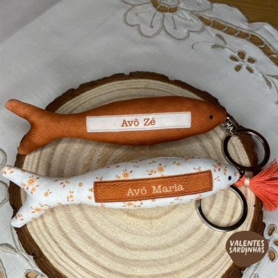 Chaveiros em forma de sardinha com etiquetas 'Avô Zé' e 'Avó Maria' sobre madeira e tecido bordado